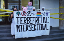 Fünf Aktivis halten ein weißes Banner mit der Aufschrift Tierbefreiung Intersektional. Auf diesem sind Symbole vom Hambi, Gegen-Nazis, Anarchismus, Tierbefreiung, Regenbogenfahne, Hausbesetzungs-N und Queerfeminismus.