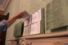 Man sieht eine Hand, die sich ein Papier von einem an die Wand gehangenen Papierstapel nimmt.