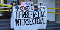 Fünf Aktivis halten ein weißes Banner mit der Aufschrift Tierbefreiung Intersektional. Auf diesem sind Symbole vom Hambi, Gegen-Nazis, Anarchismus, Tierbefreiung, Regenbogenfahne, Hausbesetzungs-N und Queerfeminismus.