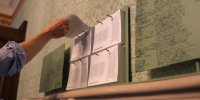 Man sieht eine Hand, die sich ein Papier von einem an die Wand gehangenen Papierstapel nimmt.